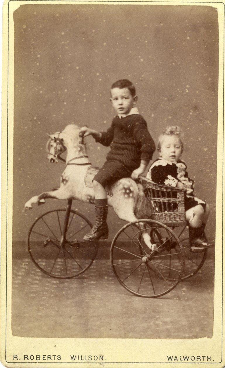 Ragazzo su cavallo a dondolo con ragazza in carrozza di vimini dietro, 1880s (stampa all